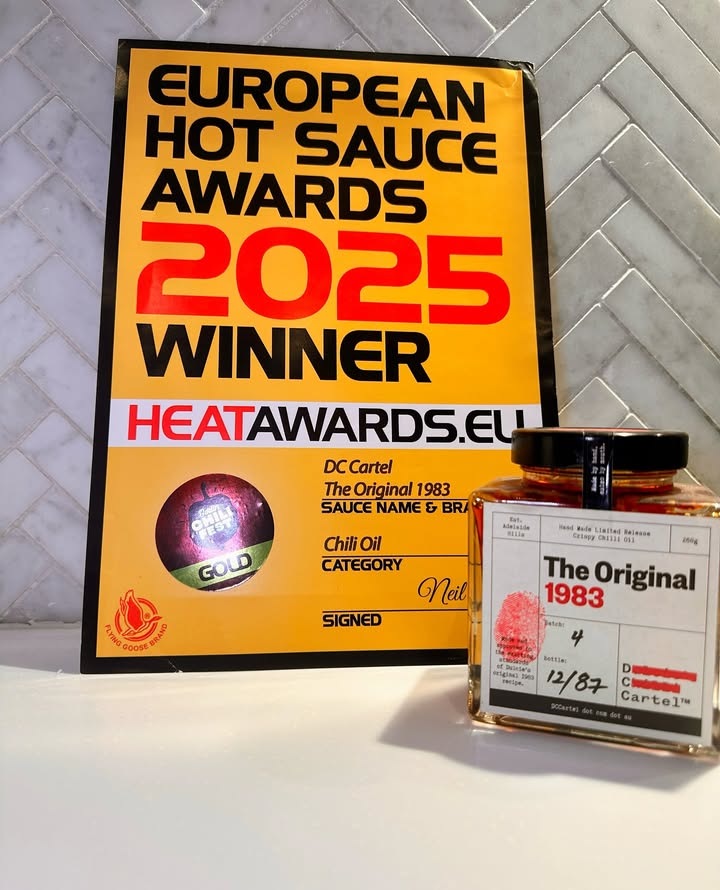 Ein Zertifikat der European Hot Sauce Awards 2025, das DC CARTEL The Original 1983 als Gewinner in der Kategorie Chiliöl auszeichnet. Daneben steht die preisgekrönte Glasflasche mit dem markanten roten Fingerabdruck.