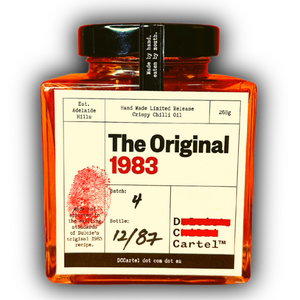 Nahaufnahme von DC CARTEL Chiliöl The Original 1983. Auf dem cremefarbenen Etikett sticht ein großer, roter Fingerabdruck hervor, der das handgemachte Design unterstreicht. Die quadratische Glasflasche ist mit leuchtend rotem Öl gefüllt. Ein Symbol für authentisches, handgefertigtes Crispy Chili Oil.